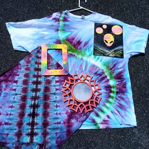 May include: A tie-dye t-shirt with an alien graphic, a tie-dye scarf, a square frame, and a decorative mirror. The t-shirt is light blue, purple, and green. The scarf is purple and blue. The frame is pink, yellow, and green. The mirror is orange.