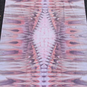 May include: A tie-dye textile with a symmetrical pattern in shades of pink, purple, and white. The design features a central diamond shape with radiating lines, creating a textured appearance. The fabric is laid flat, showcasing the intricate dye work.
