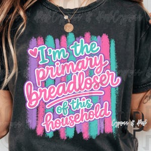 Op de afbeelding: Een zwart mineraal gewassen t-shirt met een afbeelding van verticale penseelstreken in groenblauw, paars en roze. De tekst "I'm the primary breadloser of this household" staat in een gestileerd roze en groenblauw lettertype, met een klein roze hartje.