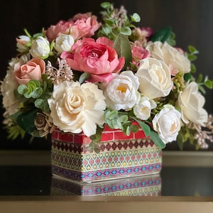 May include: A floral arrangement in a colorful patterned square vase. The arrangement features white, pink, and cream roses, along with greenery.