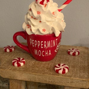 May include: A red mug with white lettering that reads "Peppermint Mocha" is filled with white whipped cream and topped with a red and white peppermint candy. A red and white striped straw is sticking out of the whipped cream. The mug is sitting on a wooden stool with four red and white peppermint candies scattered around it.