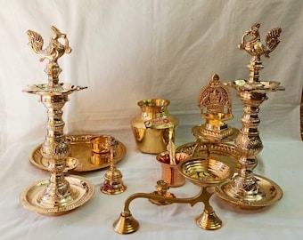 Può includere: Un set di articoli tradizionali indiani per la puja in ottone, tra cui due lampade ad olio ornate con capitelli a forma di pavone, una piccola campana, una brocca per l'acqua, un piatto e un supporto decorativo con un piccolo diya. Tutti gli articoli sono lucidati a una finitura dorata lucida.