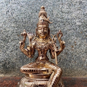 May include: A gold-colored statue of a Hindu deity, sitting in a meditative pose with four arms. The deity is adorned with intricate jewelry and a crown. The statue is set on a circular base with decorative carvings.