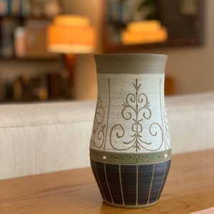 May include: A white ceramic vase with a brown rim and a black and green geometric design. The vase has a floral pattern in brown.