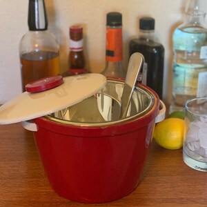 May include: A red ice bucket with a cream-colored lid and a stainless steel interior. A stainless steel ice spoon rests inside. Bottles of liquor and a glass of ice are in the background. A lemon and lime are on the right.