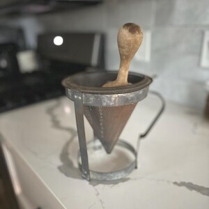 May include: A vintage metal food mill with a wooden pestle. The mill has a cone-shaped strainer and a sturdy metal frame. The wooden pestle is light brown. This kitchen tool is designed for straining and pureeing food.
