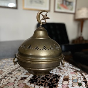 May include: A vintage brass incense burner with a crescent moon and star finial. The round body features decorative cutouts and small handles. The antique-style burner is a decorative item.