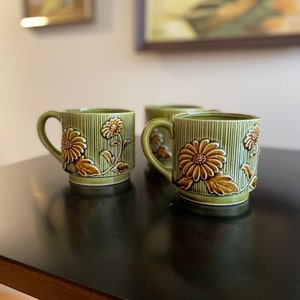 May include: Set of four green ceramic mugs with brown floral designs. The mugs have a striped pattern and are perfect for a morning cup of coffee or tea.