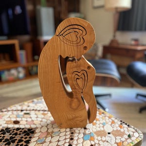 May include: Wooden sculpture of two figures embracing, carved with intricate heart shapes. The sculpture is made of light brown wood and has a natural finish.