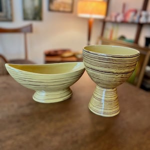 May include: Two ceramic bowls with a yellow and brown striped pattern. The bowl on the left is a shallow, oval-shaped bowl with a pedestal base. The bowl on the right is a taller, round bowl with a pedestal base.