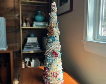 May include: A cone-shaped Christmas tree decoration covered in white pearls, blue, red, and gold beads. The tree is about 12 inches tall and is sitting on a wooden table.
