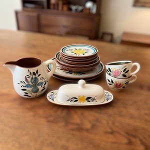 May include: A set of vintage floral patterned ceramic dishes, including a creamer, butter dish, and two teacups. The dishes feature a variety of floral designs, including irises, tulips, and daisies, in shades of blue, yellow, pink, and green.