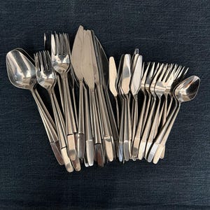 May include: A collection of stainless steel silverware, including spoons, forks, and knives, arranged on a dark blue textured surface. The silverware has a simple, elegant design, reflecting light. The image showcases a complete set of dining utensils.