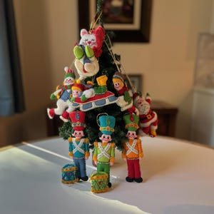 May include: A collection of felt Christmas ornaments, including a white bunny, a snowman, a sleigh with three figures, a Santa Claus, and three toy soldiers. The ornaments are decorated with colorful beads and sequins.