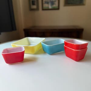 May include: A collection of colorful, rectangular glass baking dishes. The dishes are in various colors, including red, yellow, and blue. The dishes are arranged on a white surface. The interior of the dishes is white.