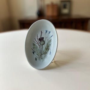 May include: A small, oval-shaped ceramic dish with a blue and green floral design featuring a thistle. The dish is white with a light blue glaze.