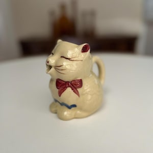 May include: A cream-colored ceramic cat creamer with a red bow and blue accents. The cat has a playful expression and is sitting on its hind legs.