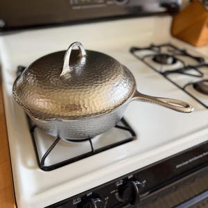 May include: A hammered metal skillet with a lid, sitting on a white stovetop. The skillet has a long handle and a textured surface. The lid has a curved handle. The stovetop has black burners and control knobs.