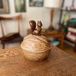 May include: A ceramic nutcracker shaped like a walnut with a squirrel figurine on top. The nutcracker is brown and beige with a detailed texture.