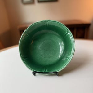 May include: A green ceramic bowl with a scalloped edge, resting on a black metal stand. The bowl has a glossy finish and a deep, rich green color. The bowl is round and appears to be empty.