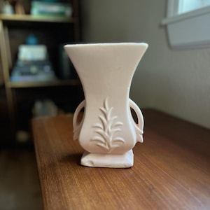 May include: A pale pink ceramic vase with a flared top and a square base. The vase features two small handles and a raised leaf design on the front. The vase is sitting on a wooden surface.