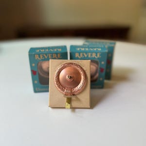 May include: A copper-colored Revere picture hanger with a decorative circular design. The hanger is mounted on a small cardboard backing, with the word "REVERE" visible on the blue boxes in the background.