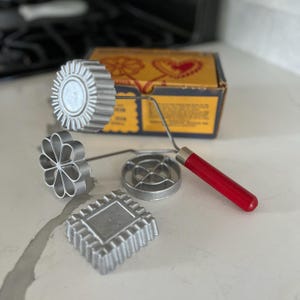 May include: Set of four vintage metal cookie cutters with a red handle. The cutters are shaped like a flower, a circle, a square, and a sun. The cutters are in a box with a yellow and red design.