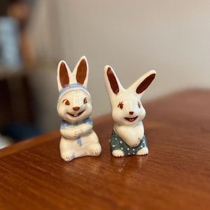 May include: Two white ceramic bunny figurines with brown ears. One bunny is wearing a blue and white scarf and the other is wearing green and white pants.