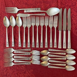 May include: A set of silver flatware, including forks, knives, and spoons, arranged in a row on a red tablecloth. The flatware has ornate handles with decorative designs.