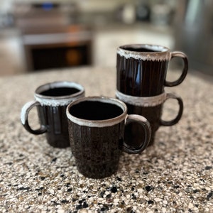 May include: Four brown ceramic mugs with white rims. The mugs have a textured, almost woven, pattern. The mugs are stacked on top of each other.
