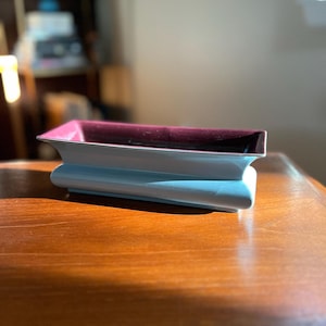 May include: A rectangular ceramic planter with a light blue exterior and a dark purple interior. The planter has a slightly rounded shape and a smooth finish.