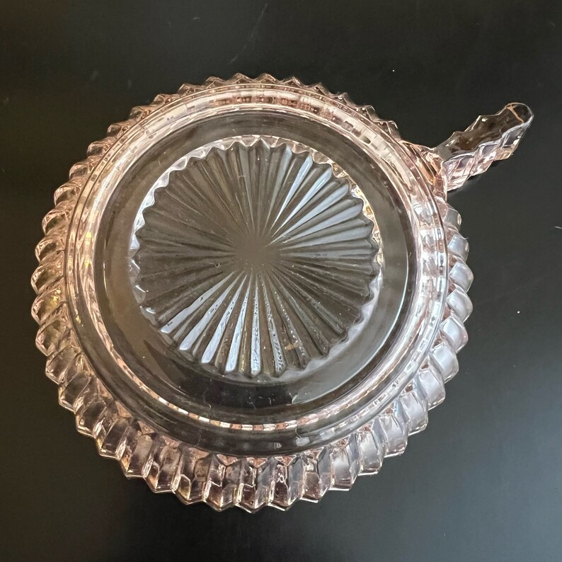 Puede incluir: Un plato de vidrio rosa con un borde festoneado y un asa. El plato tiene un patr&oacute;n de rayos de sol en el centro.