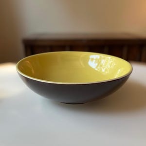 May include: A yellow and brown ceramic bowl. The bowl is round and has a wide rim. The inside of the bowl is yellow and the outside is brown.