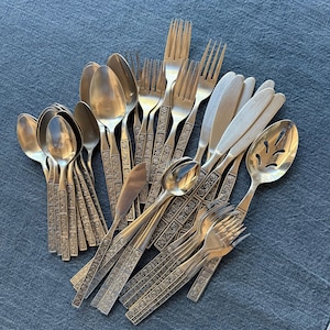 May include: A collection of stainless steel flatware featuring a detailed floral pattern on the handles. The set includes various forks, spoons, knives, and a slotted serving spoon, all arranged in a cluster with polished metal surfaces.