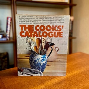 May include: The cover of a vintage cookbook titled "The Cooks' Catalogue". The cover features a blue and white enamel pitcher filled with kitchen utensils, including whisks, spoons, and a metal spatula. The pitcher is sitting on a wooden table with a brown earthenware pitcher behind it.