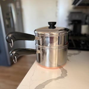 May include: A stainless steel double boiler pot with a matching lid. The unit features two stacked cylindrical sections, each equipped with a black heat-resistant handle containing a metal hanging loop. The base of the bottom pot has a copper finish.