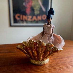 May include: A gold ceramic seashell-shaped dish with a doll wearing a pink dress sitting inside. The doll has black hair and brown eyes.