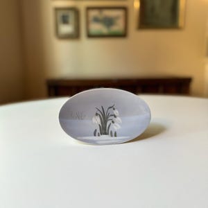 May include: Oval-shaped decorative dish with a light blue background. The dish features a painted scene of snowdrops in a snowy landscape. The snowdrops are white with green stems and leaves.