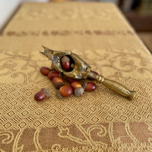May include: A gold-toned nutcracker shaped like a hand, with a nut in the palm, surrounded by acorns on a brown patterned fabric.
