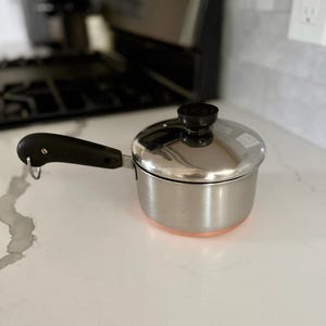 May include: A small, stainless steel saucepan with a lid and a black handle. The pot has a copper-colored bottom and sits on a white countertop. The lid has a black knob. The handle has a loop for hanging.