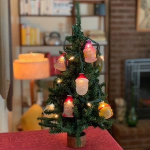 May include: A small artificial Christmas tree decorated with string lights and glass ornaments shaped like faces. The ornaments are in shades of pink, red, and white.
