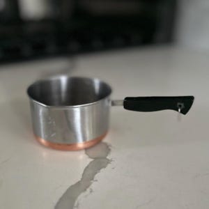 May include: A small stainless steel saucepan with a copper-colored base and a black handle. The pan is sitting on a white countertop. The handle is attached with a small metal rivet.