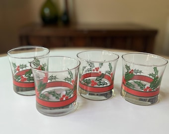 Op de afbeelding: Vier heldere drinkglazen met een feestelijk kerstontwerp. Elk glas heeft een rode band met hulst- en bessenaccenten. De glazen zijn gerangschikt op een wit oppervlak.