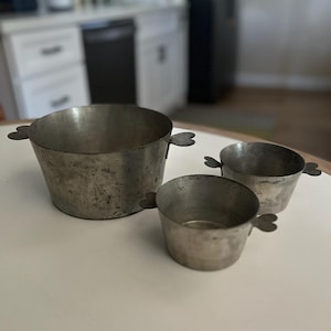 May include: Three silver metal measuring cups with heart-shaped handles. The cups are nested together, with the largest cup at the bottom and the smallest cup at the top.