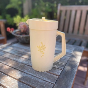 May include: A white pitcher with a yellow floral design and a yellow lid. The pitcher is sitting on a wooden table.