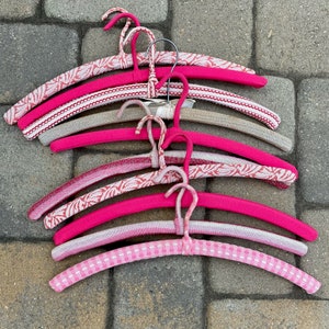 Puede incluir: Una pila de seis perchas de color rosa y blanco con un gancho de alambre. Las perchas est&aacute;n hechas de un material suave y flexible, ideales para colgar art&iacute;culos delicados.