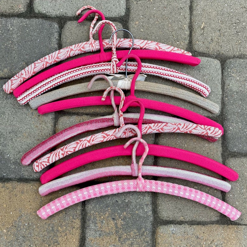 Puede incluir: Una pila de seis perchas de color rosa y blanco con un gancho de alambre. Las perchas est&aacute;n hechas de un material suave y flexible, ideales para colgar art&iacute;culos delicados.