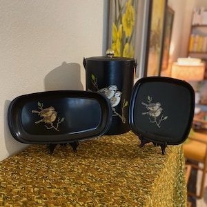 May include: Three black trays with gold bird designs. The trays are on small black stands. The trays are oval, square, and round. The round tray is a vintage ice bucket.