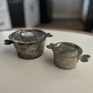 May include: Two vintage silver-colored metal pots with lids and handles. The larger pot has a brown handle and the smaller pot has a silver handle. Both pots have a slightly distressed finish.