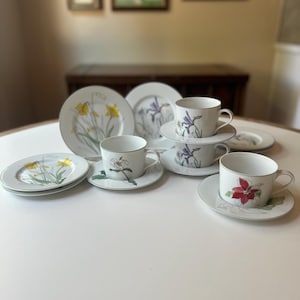 May include: A collection of fine china with floral designs. The set includes plates, saucers, and teacups, each adorned with botanical illustrations of daffodils, irises, and other flowers. The rims are trimmed with a delicate green border.